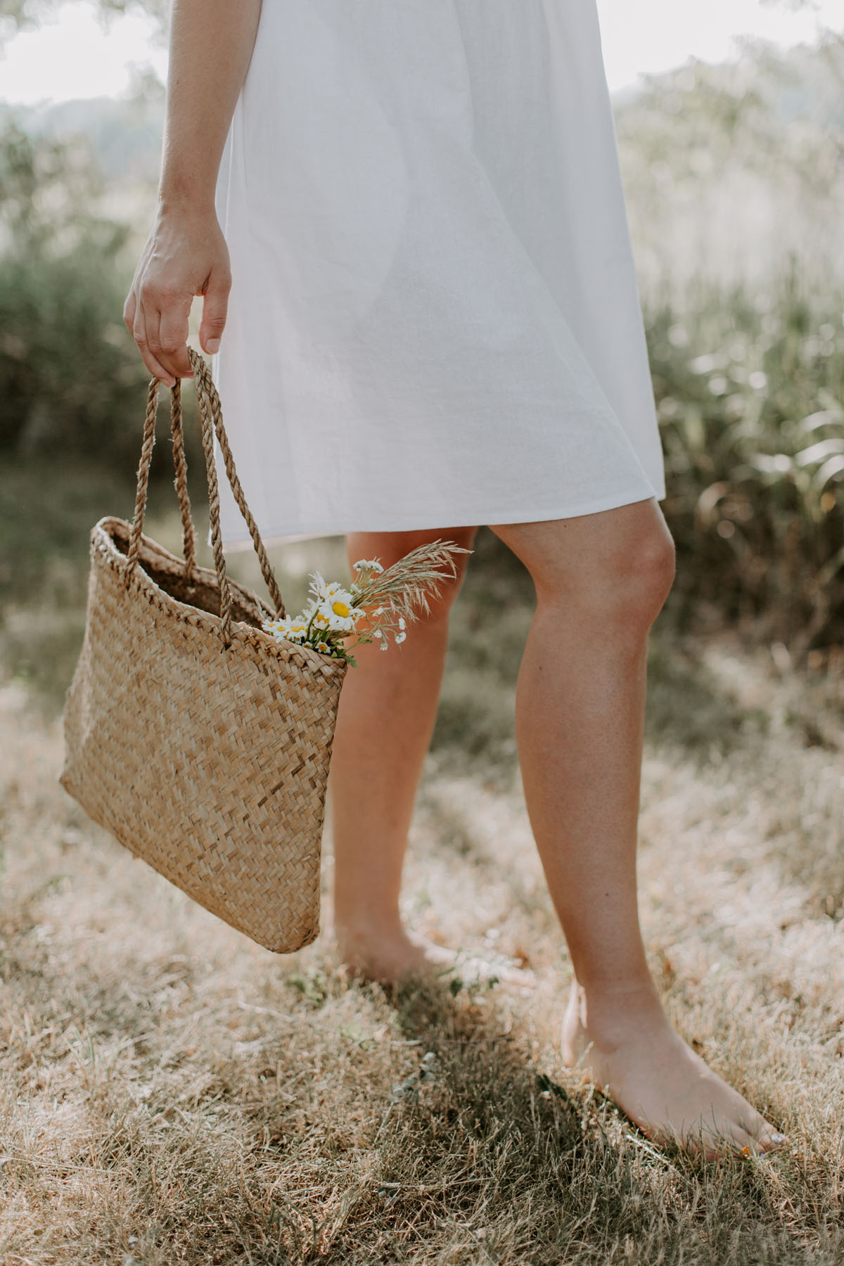 image shows a woman wearing a white dress and holding a woven bag
